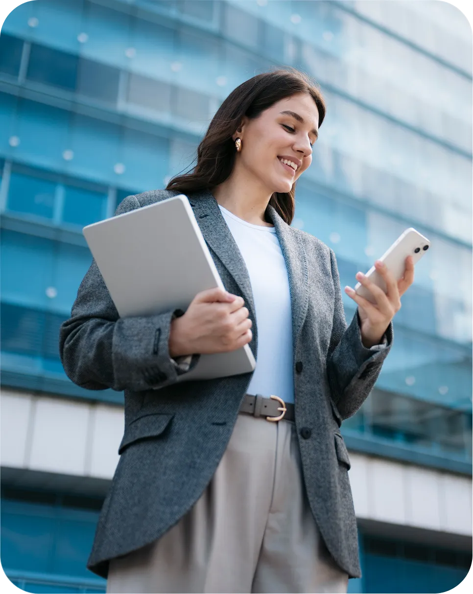 Professional holding a laptop and reviewing information on a smartphone outside an office building